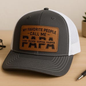 May include: A grey and white trucker hat with a brown leather patch. The patch reads "MY FAVORITE PEOPLE - CALL ME - PAPA" with the names Liam, Oliver, Sophia, and Harper below. A black pen and glasses are also in the image.