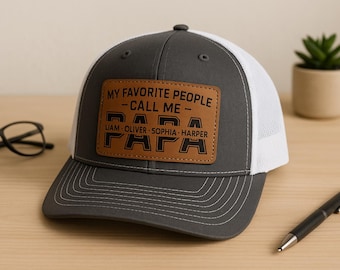 Könnte beinhalten: Grau-weiße Trucker-Mütze mit braunem Lederaufnäher. Der Aufnäher lautet "MY FAVORITE PEOPLE - CALL ME - PAPA" mit den Namen Liam, Oliver, Sophia und Harper darunter. Ein schwarzer Stift und eine Brille sind ebenfalls im Bild.