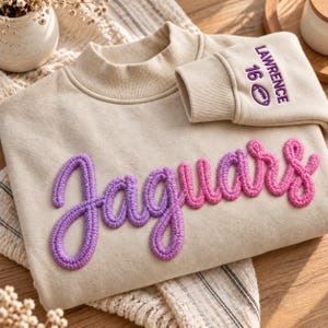 Könnte beinhalten: Ein beigefarbenes Sweatshirt mit dem Wort "jaguars" in einer violett-rosa verlaufenden Chenille-Schrift auf der Brust. Der Ärmelbund ist mit dem Text "LAWRENCE 16" und einem kleinen Football-Symbol in violettem Garn bestickt.