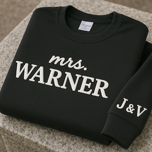 May include: Black sweatshirt with the text "mrs. WARNER" in white block letters across the front. The sleeve has the initials "J&V" in white. The Gildan brand label is visible.