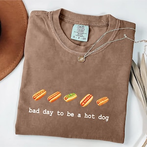 Pode incluir: Camiseta marrom com a frase "bad day to be a hot dog" e um gráfico de cinco cachorros-quentes. A camiseta tem gola redonda e mangas curtas. Um colar de prata com um pequeno pingente está na camiseta.