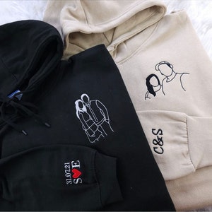 May include: Three hooded sweatshirts in black and beige. Each sweatshirt features custom line art embroidery of a couple on the front. One black sweatshirt has the date "31.07.21" and initials "S E" embroidered on the sleeve. The beige sweatshirt has the initials "C & S" on the sleeve.