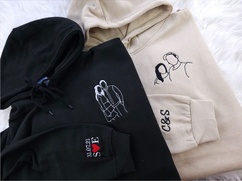 May include: Three hooded sweatshirts in black and beige. Each sweatshirt features custom line art embroidery of a couple on the front. One black sweatshirt has the date "31.07.21" and initials "S E" embroidered on the sleeve. The beige sweatshirt has the initials "C & S" on the sleeve.