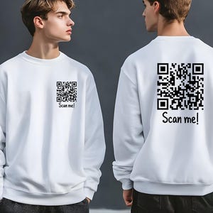 May include: A white long-sleeve sweatshirt featuring a black QR code graphic on the left chest and a larger matching QR code graphic on the back. Both graphics include the text "Scan me!" printed in a black sans-serif font below the code.