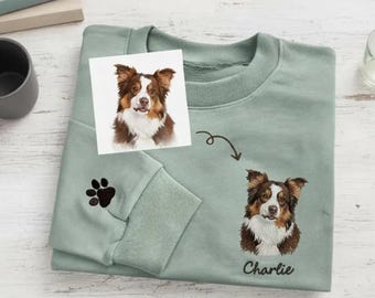 May include: A light green sweatshirt featuring a custom dog portrait and paw print. The sweatshirt has a watercolor portrait of a dog on a small card and an embroidered dog portrait with the name "Charlie" below it.