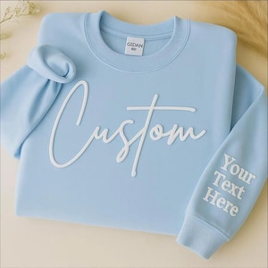 May include: A light blue sweatshirt with the word "Custom" in white script across the front. The right sleeve has the text "Your Text Here" in white. The sweatshirt has a crew neck and ribbed cuffs.