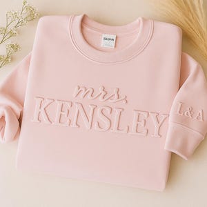 May include: A light pink sweatshirt with the words "mrs. KENSLEY" in raised lettering across the front. The sleeve cuff has the initials "L&A". The Gildan brand tag is visible at the neckline. The sweatshirt is a crew neck style.