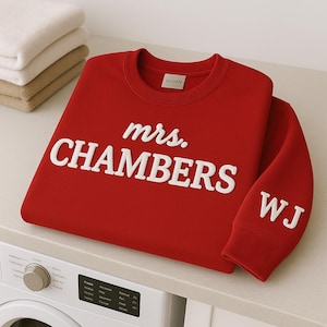 May include: A red sweatshirt with the text "mrs. CHAMBERS" in white block letters across the front. The sleeve has the initials "WJ" in white. The sweatshirt is folded on a light-colored surface.