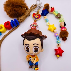 Op de afbeelding: Een sleutelhanger met een Woody-figuurtje met een bruine cowboyhoed, geel overhemd en blauwe broek. Het bevat een bruine imitatiebont pompon, een beige koord en kleurrijke kralen die woorden spellen. Een hartvormige bedel met een afbeelding is bevestigd.