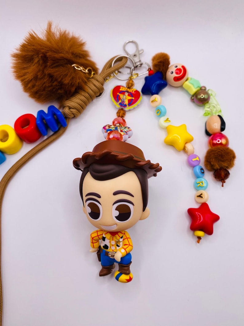 Op de afbeelding: Een sleutelhanger met een Woody-figuurtje met een bruine cowboyhoed, geel overhemd en blauwe broek. Het bevat een bruine imitatiebont pompon, een beige koord en kleurrijke kralen die woorden spellen. Een hartvormige bedel met een afbeelding is bevestigd.