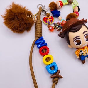Op de afbeelding: Een sleutelhanger met een bruin paracord koord waarop de naam WOODY is gespeld met kleurrijke plastic letterkralen. Het bevat een bruine imitatiebont pompon, een klein Woody-figuurtje en diverse decoratieve kralen, waaronder een sheriffster-bedel.