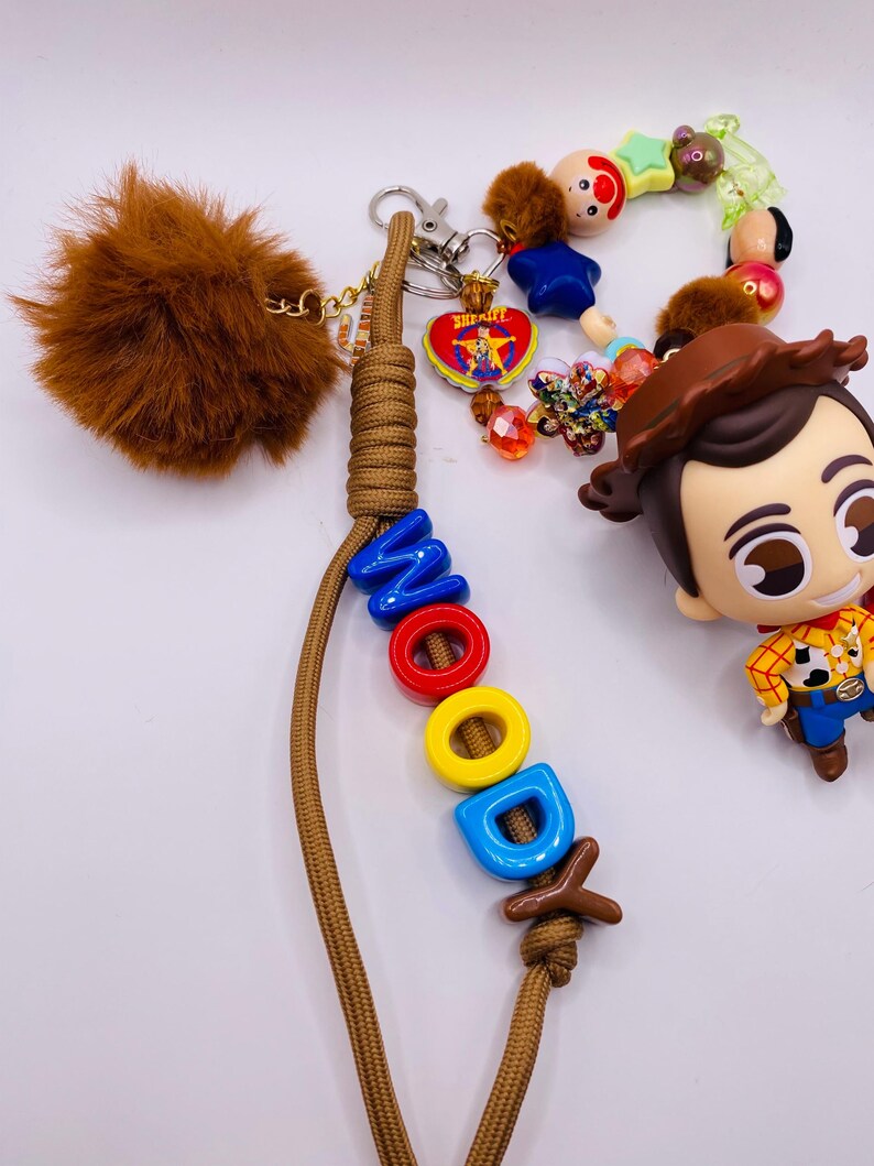 Op de afbeelding: Een sleutelhanger met een bruin paracord koord waarop de naam WOODY is gespeld met kleurrijke plastic letterkralen. Het bevat een bruine imitatiebont pompon, een klein Woody-figuurtje en diverse decoratieve kralen, waaronder een sheriffster-bedel.