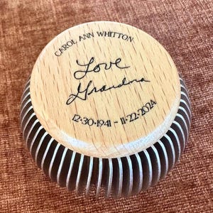 May include: A wooden-topped container with the inscription "CAROL ANN WHITTON" and "Love Grandma" in cursive. The dates "12-30-1941 - 11-22-2024" are also engraved. The container has a ribbed, silver and black design.