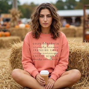 May include: A coral-colored long-sleeve sweatshirt featuring a large yellow maple leaf graphic printed on the center chest. The text reads: cozy blankets, crunchy leaves, crackling fires, crisp mornings, chilly nights, comfy sweaters.