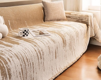 May include: A beige sofa with a cream-colored throw blanket featuring a brushstroke design. A matching pillow with a line drawing rests on the sofa. A black and white checkered mug and a magazine are on the sofa.