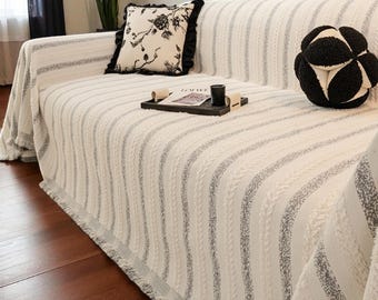 May include: A white sofa blanket with vertical gray stripes and a fringed edge. The sofa is adorned with decorative pillows, including one with a floral design and another in a black and white geometric pattern.