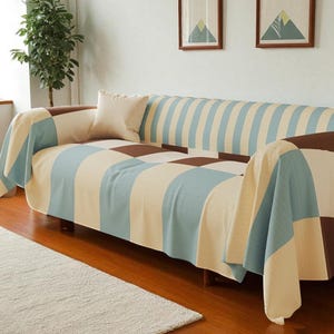 Puede incluir: Un sofá cubierto con una manta estampada en azul, marrón y crema. La manta tiene un diseño de tablero de ajedrez en el asiento y un patrón de rayas en el respaldo. Una almohada color crema descansa sobre el sofá.