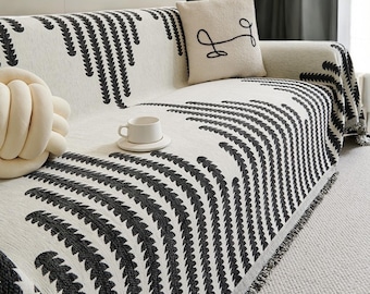 May include: A white sofa with a black and white patterned throw blanket. The throw blanket has a fringe border and a geometric design. A white throw pillow with black embroidery is on the sofa.