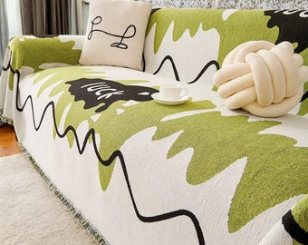 May include: A white and green sofa throw with a black and white abstract design. The throw has a fringe border and features the word "luck" in black lettering. A white knotted pillow is on the sofa.