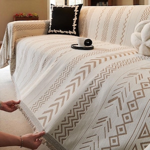 Puede incluir: Funda de sofá color crema con un estampado geométrico en tonos marrones. La funda tiene flecos en el borde y está colocada sobre un sofá. También se ve un cojín negro con borde blanco y un cojín decorativo blanco.