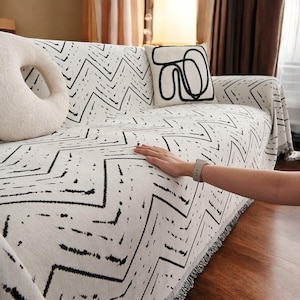 May include: A white and black geometric patterned throw blanket with fringe on the edges. The blanket is draped over a sofa with a white throw pillow with a black abstract design.