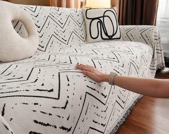May include: A white and black geometric patterned throw blanket with fringe on the edges. The blanket is draped over a couch with a white throw pillow with a black abstract design.