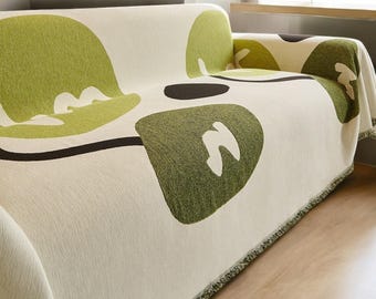 May include: A white sofa throw blanket with a green and brown abstract design. The blanket has a fringe border.