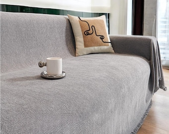 May include: A gray sofa throw blanket with a fringe border. The blanket is draped over a sofa with a decorative pillow. A white ceramic cup and saucer are on the sofa.