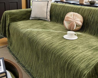 May include: A green textured sofa cover with a fringe trim, draped over a sofa. A beige pillow with a black border and a decorative round pillow with a flower design are on the sofa. A white teacup and saucer are also visible.