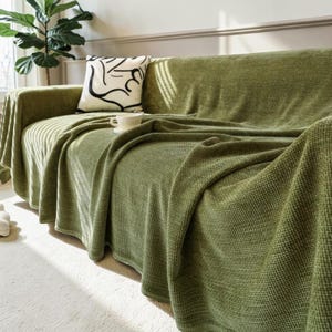May include: A sage green textured throw blanket draped over a sofa, featuring a waffle-knit pattern. A white throw pillow with a black abstract line drawing sits on the sofa, accompanied by a small white ceramic cup placed on the blanket surface.