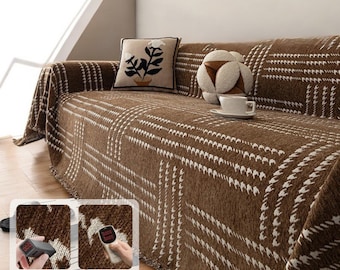 May include: A brown and white patterned throw blanket with fringe edges draped over a couch. The blanket has a geometric design with white squares on a brown background. The couch has a white pillow with a black floral design and a brown and white round pillow.