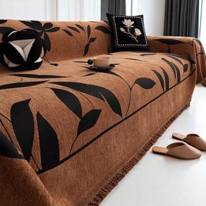 Peut inclure: Une housse de canapé marron avec un motif de feuilles noires. La housse est posée sur un canapé, avec une boule décorative noire et blanche et un coussin noir avec une broderie florale blanche. Une tasse et une soucoupe sont sur le canapé, et des pantoufles marron sont au sol.