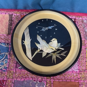 Puede incluir: Reloj de pared redondo con esfera negra, con un diseño de pájaros y bambú. El reloj tiene un borde color crema y un borde exterior negro. Los pájaros y el bambú están hechos de un material nacarado. Las manecillas del reloj son plateadas.