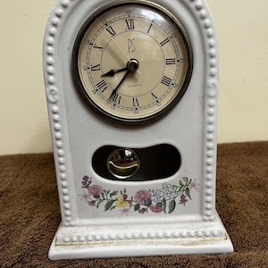 May include: White ceramic quartz clock with a decorative floral design and a swinging pendulum. The clock face has Roman numerals and the letters "PS". The clock is approximately 18 cm tall.