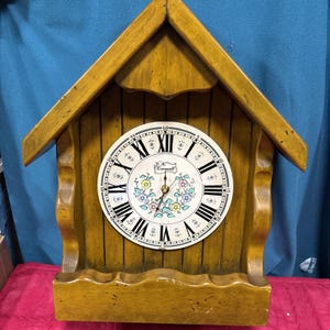 May include: A wooden wall clock with a house-shaped frame. The clock face is white with black Roman numerals and a floral design in the center. The clock's frame is a light brown color.