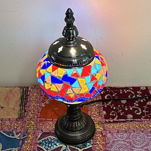 May include: A table lamp featuring a spherical glass shade covered in a mosaic of triangular red, blue, yellow, and light blue glass pieces. The lamp has a dark bronze-colored metal base and a matching metal cap with a decorative finial top.