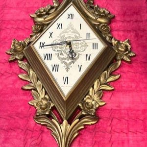 May include: A gold-toned wall clock with a diamond-shaped face, Roman numerals, and ornate floral and leaf detailing. The clock has a decorative bow at the top and a stylized floral base. The background is a pink fabric.