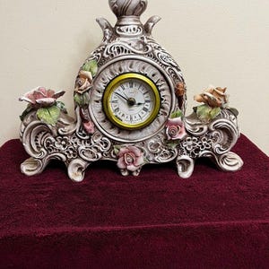 Puede incluir: Un reloj de cerámica decorativo para repisa con acabado blanco y gris, adornado con detalles florales. La esfera del reloj tiene un bisel dorado y números romanos negros. El reloj mide aproximadamente 25 cm de ancho.