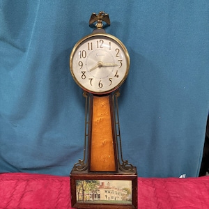 May include: Antique Sessions clock with a gold-tone eagle finial. The clock face has black numerals and hands, with a gold-tone bezel. The wooden body features a burl wood panel and a framed picture of a building at the base. The clock is approximately 18 inches tall.