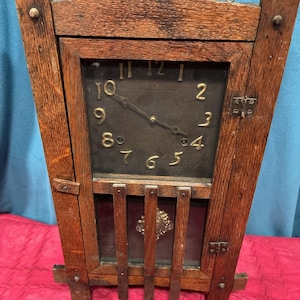 May include: Antique wooden clock with a dark face and visible clock hands. The clock has a rectangular shape with a decorative wooden frame. The clock face features the numbers 1 through 12. The clock is a vintage home decor item.