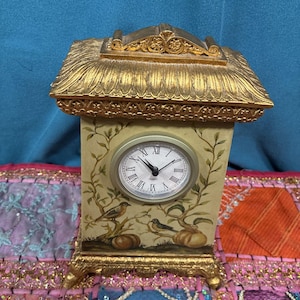 May include: A decorative quartz clock with a gold-colored roof and base. The clock face has Roman numerals and black hands. The clock's body is painted with birds and vines. The clock is a decorative home accent.