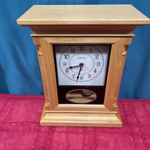 Könnte beinhalten: Eine Hampton-Uhr aus Holz mit hellbraunem Finish. Das Zifferblatt ist weiß mit schwarzen Ziffern und Zeigern. Ein kleines Pendel schwingt unterhalb des Zifferblatts. Die Uhr ist etwa 30 cm hoch.