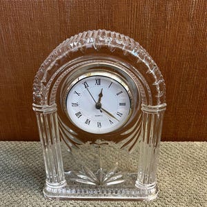 Puede incluir: Un reloj de cuarzo de cristal transparente con esfera blanca y números romanos negros. El reloj tiene una parte superior y una base arqueadas decorativas con un diseño floral. El reloj mide aproximadamente 15 cm de altura.