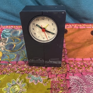 May include: A dark blue, rectangular analog clock with a white face, black numbers, and black hands. The second hand is red, and the clock has a musical note design on the bottom. The clock is on a colorful patterned surface.