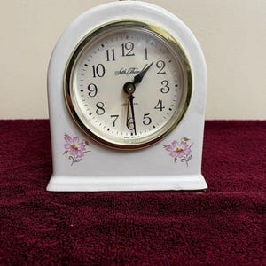 May include: White Seth Thomas alarm clock with a gold-tone bezel and a light dial. The clock has a curved top and floral accents. The clock face displays black numerals and hands. The clock is made in the USA.