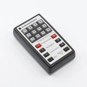 Könnte beinhalten: Eine schwarze Fernbedienung mit silbernem Gesicht und Grundig-Logo. Die Fernbedienung hat ein Ziffernblatt, Tasten für Programm, Farbkontrast, Helligkeit, Lautstärke und Schnellton.