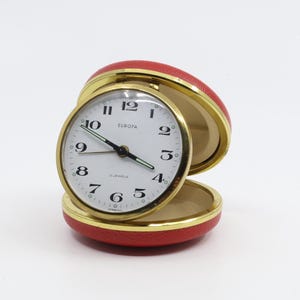 May include: A red, round, vintage alarm clock with a gold face and black hands. The clock face reads "EUROPA" and "2 JEWELS" and is set to 10:10. The clock is open and the back is visible.