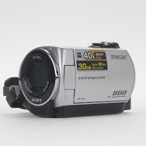 Op de afbeelding: Een zilverkleurige Sony Handycam DCR-SR32 camcorder met een zwarte lensbehuizing. De zijkant bevat een Carl Zeiss Vario-Tessar lenslabel, 40x optische zoom, 30GB harde schijfcapaciteit en tekst voor maximaal 20 uur aan opnametijd.