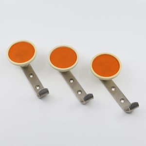 May include: Three orange and white round wall hooks with silver metal arms. The hooks are mounted on a white wall.