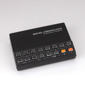 以下が含まれることがあります： IDEAL-COMPUTER PROGRAMM-CASSETTEとラベルが貼られた黒い長方形の電子制御ユニット。銀色の縁取りが施された正方形のボタンが2列に並んでおり、1から8までの番号が振られています。上段の最後の2つのボタンはオレンジ色です。